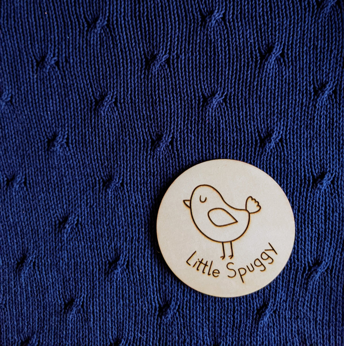 Wooden disc with a "little spuggy" picture laser etched on. Lying on a Cabled Kisses jumper